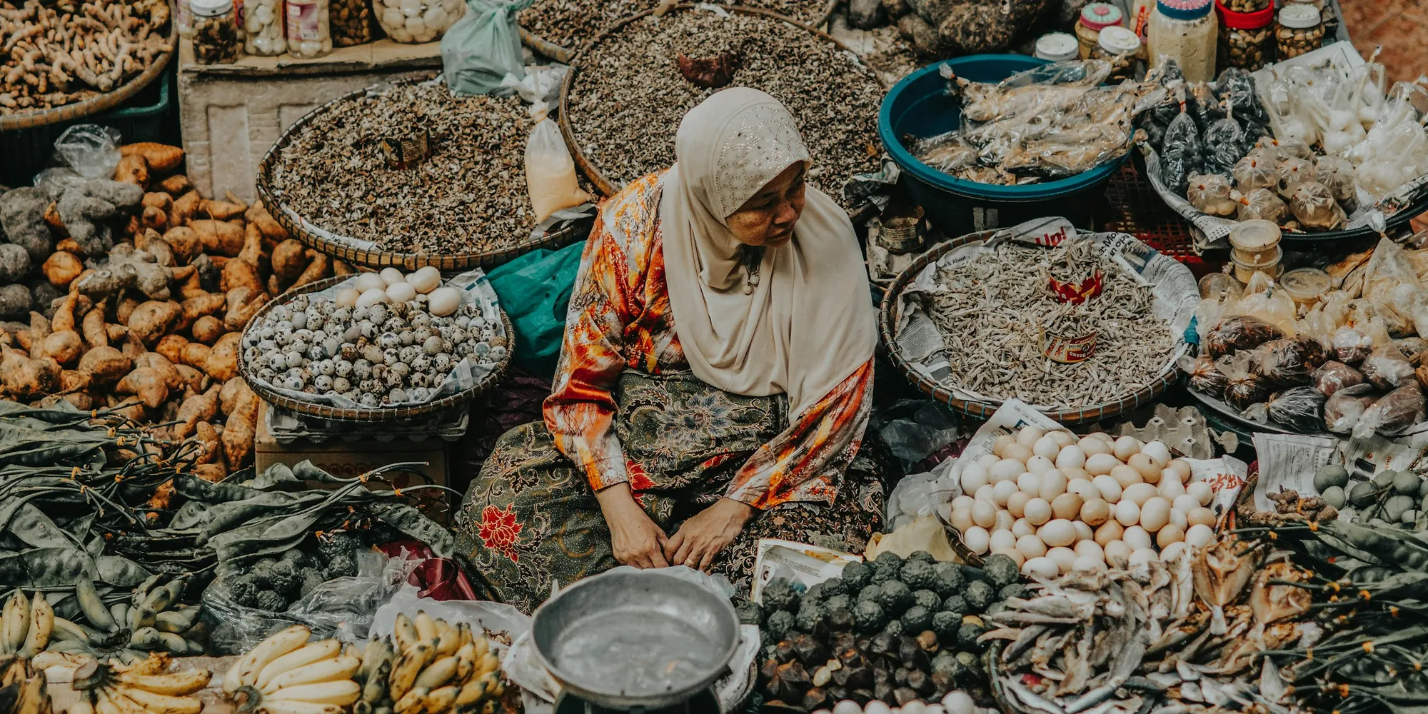 A trader in a headscarf surrounded by baskets of dried goods and produce at a Malaysian market.