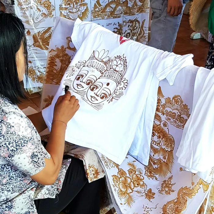 An artisan hand-drawing batik motifs onto white cloth beside finished shirts.